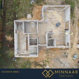 MINNAAR PARTNERS SPÓŁKA Z OGRANICZONĄ ODPOWIEDZIALNOŚCIĄ - Widok z drona na fundamenty domu z betonu, z widocznymi zbrojeniami i pracownikami w żółtych kamizelkach. Teren budowy otoczony zielenią i ziemią.