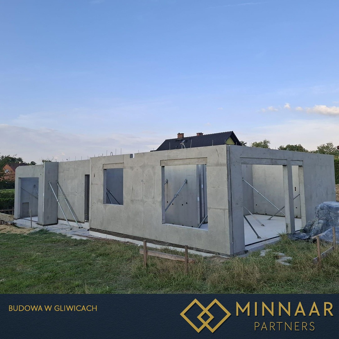 Surowy stan budynku żelbetowego z widocznymi otworami okiennymi i podporami, na tle zieleni i błękitnego nieba. Budowa w Gliwicach.