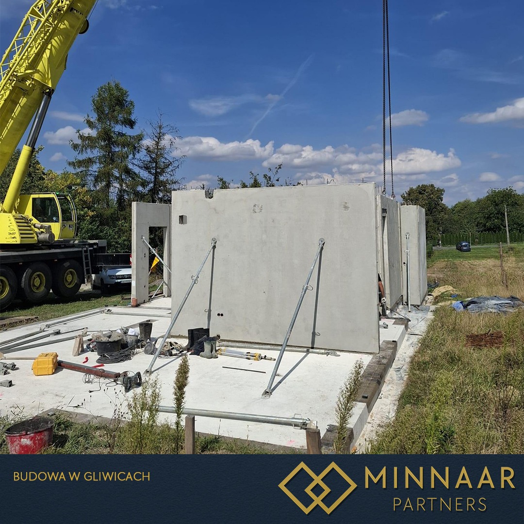 Montaż prefabrykowanych ścian betonowych na fundamencie. Żuraw budowlany w tle. Widoczne podpory stabilizujące ściany. Budowa w toku, słoneczny dzień.