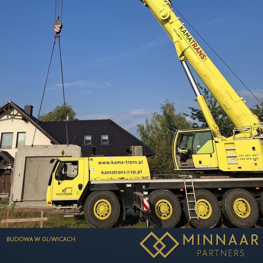 Żółty dźwig transportuje betonowy element konstrukcyjny na tle domu jednorodzinnego i błękitnego nieba. Widoczny napis 'Kamatrans' i dane kontaktowe firmy.