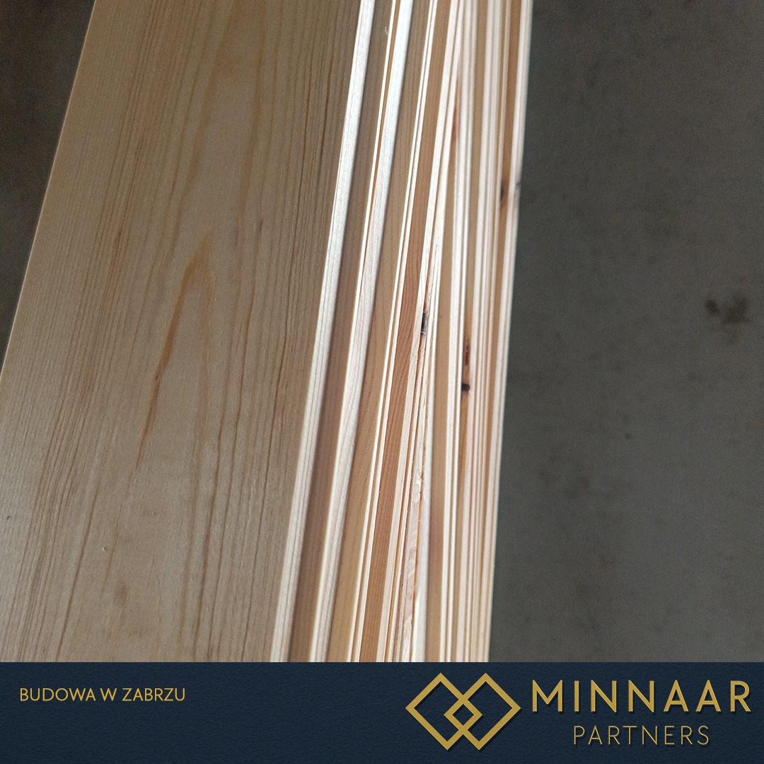 Stos jasnych, profilowanych desek drewnianych z widocznym usłojeniem, ułożonych pionowo, z logo firmy budowlanej w dolnej części kadru.