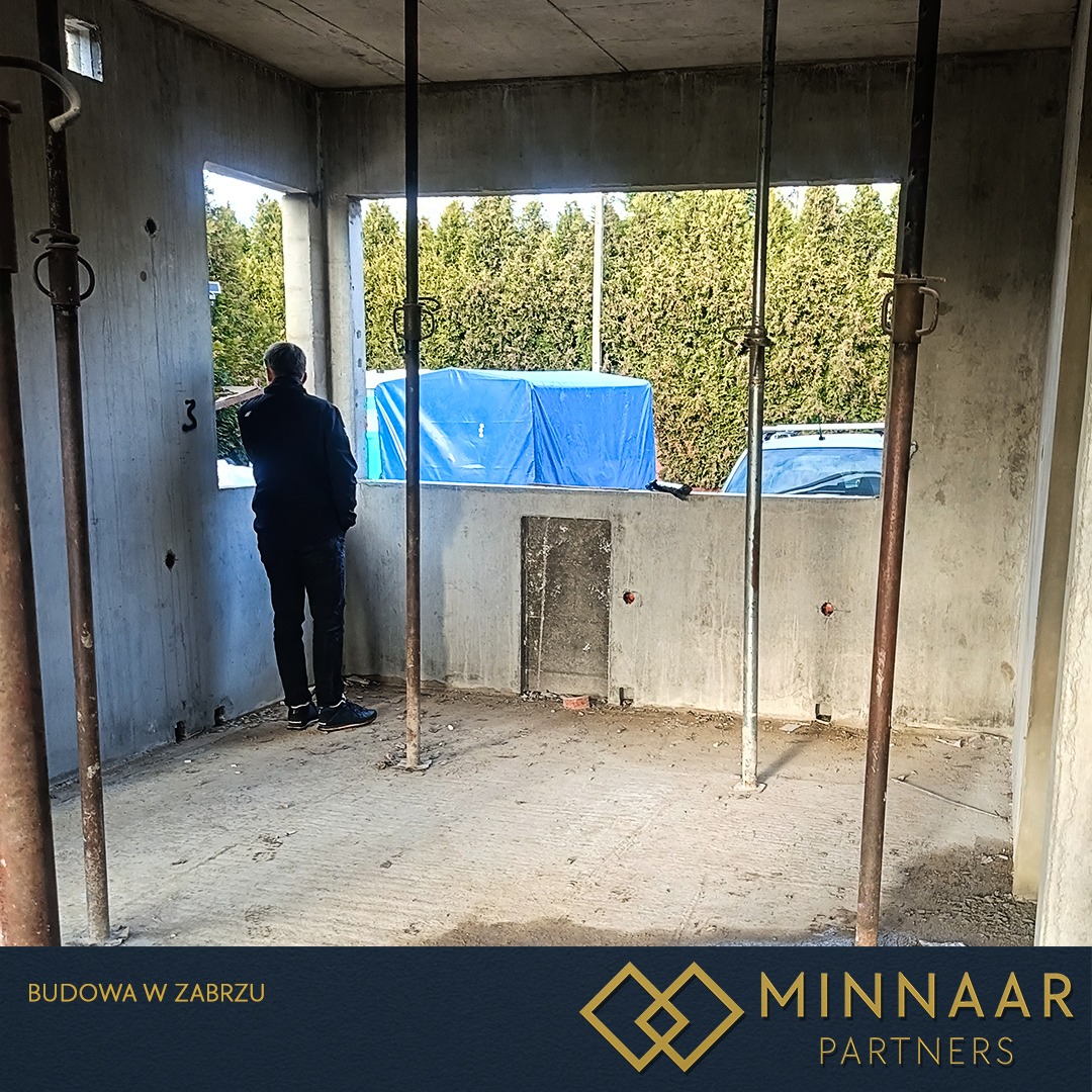 Surowy, betonowy szkielet budynku w trakcie budowy. Widoczne podpory, otwory okienne i fragment sylwetki osoby obserwującej otoczenie. W tle zieleń i niebieska plandeka.