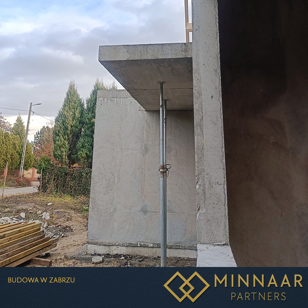 Surowa konstrukcja żelbetowa w trakcie budowy, wsparta metalowymi stemplami. Widoczne szalunki i przygotowane drewno. Ujęcie z dołu, pochmurne niebo w tle.