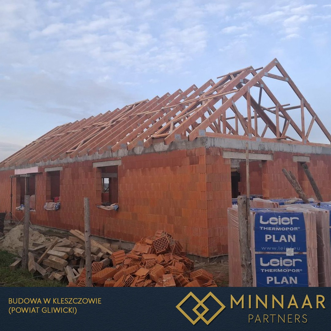 Budowa domu z czerwonej cegły: ściany wzniesione, więźba dachowa w trakcie montażu, palety cegieł Leier. Widok z zewnątrz, etap surowy otwarty.