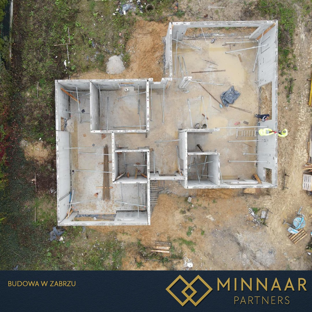 Widok z drona na fundamenty domu z betonu, z widocznymi zbrojeniami i pracownikami w żółtych kamizelkach. Teren budowy otoczony zielenią i ziemią.