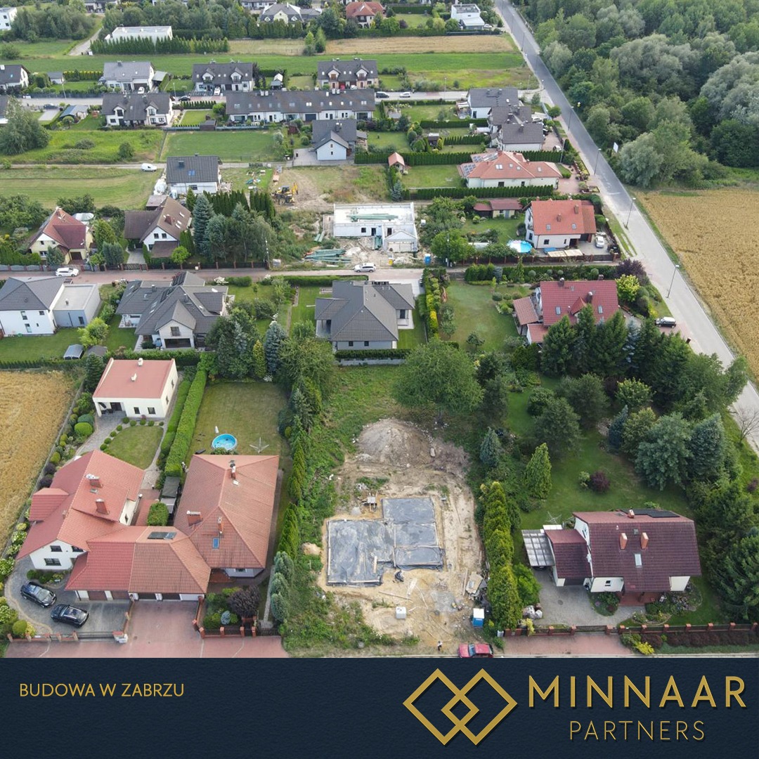 Widok z lotu ptaka na fundamenty domu jednorodzinnego w trakcie budowy, otoczone zielenią i innymi zabudowaniami. Ujęcie z drona, słoneczny dzień.