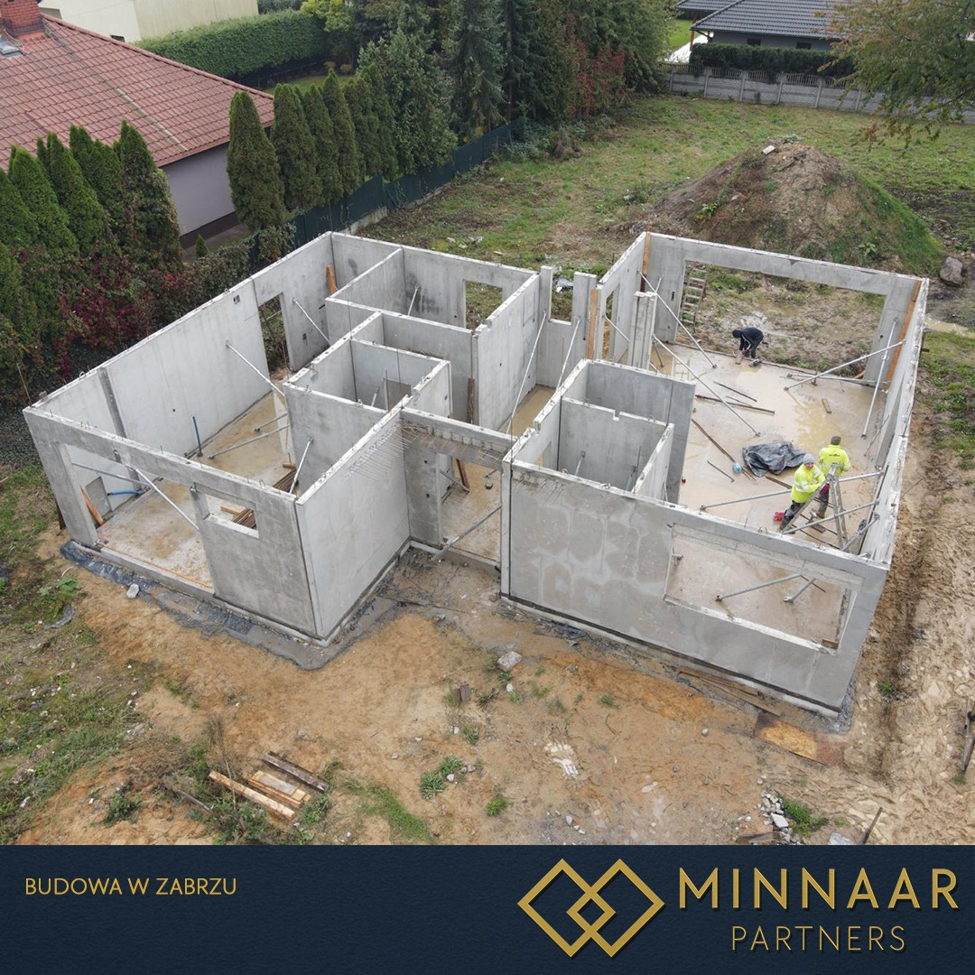 Budowa domu z betonu: widok z góry na wzniesione ściany działowe i zewnętrzne, z pracownikami w środku. W tle zieleń i domy.