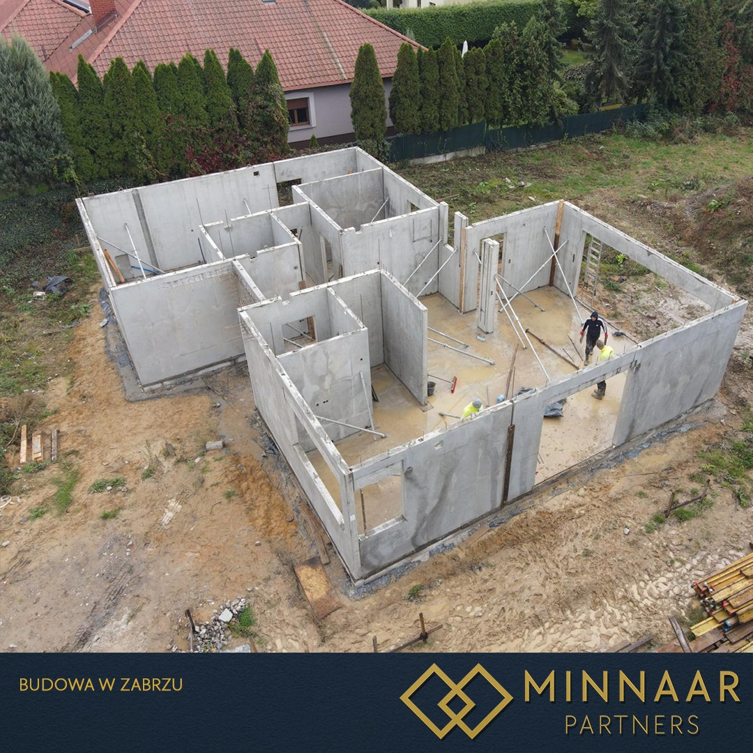 Budowa domu z prefabrykatów betonowych w toku, widok z góry. Wewnątrz pracują dwie osoby w kaskach. Na dole logo firmy budowlanej.