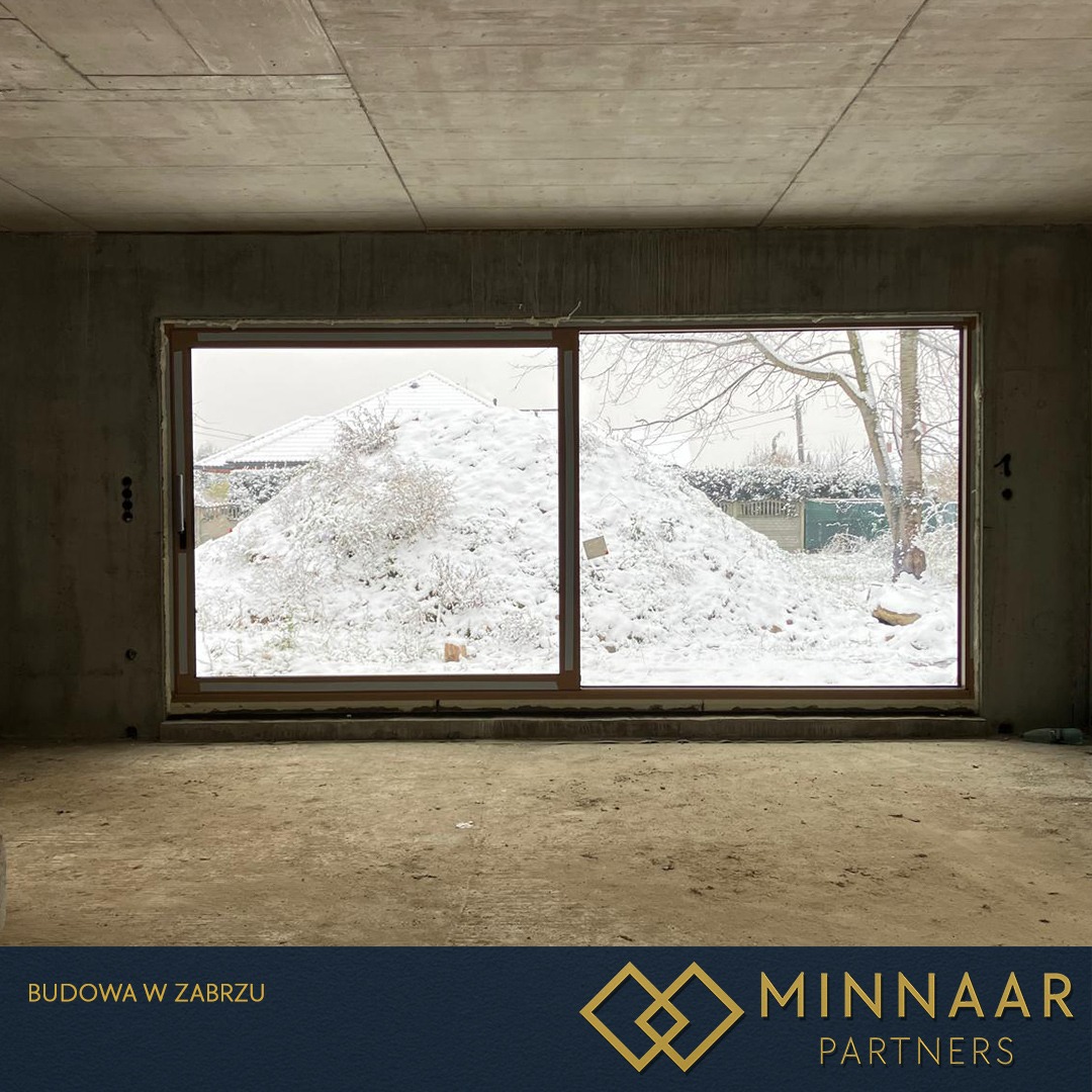 Surowy stan budynku z betonowym stropem i dużym oknem, przez które widać zasypany śniegiem ogród. Widoczny etap budowy domu w zimowej scenerii.