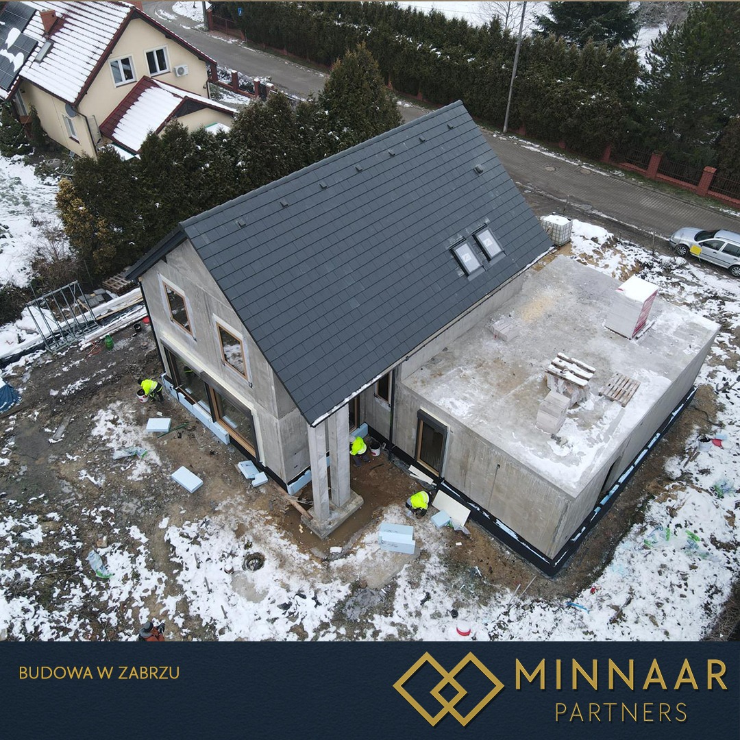 Surowy dom z szarą dachówką, widok z góry. Częściowo ocieplony fundament, okna wstawione, prace wykończeniowe w toku. Zimowy krajobraz, śnieg na ziemi.