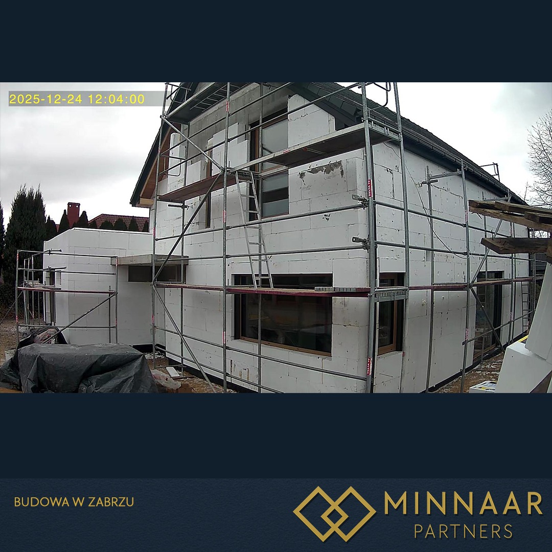 Niedokończony dom z białych bloczków, otoczony rusztowaniem. Widoczne okna i fragment dachu. Ujęcie z kamery budowlanej z datą w lewym górnym rogu.
