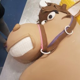 Tichy Sp.z o.o. - Uśmiechnięty tort w kształcie konia z brązowym kantarem i białymi zębami, leżący na metalowym blacie. W tle widoczne narzędzia cukiernicze.