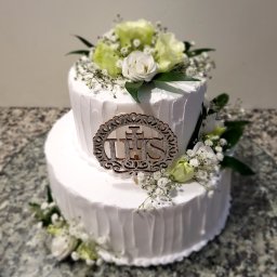 Tichy Sp.z o.o. - Dwupoziomowy tort komunijny z białym kremem, dekorowany żywymi kwiatami i srebrnym topperem IHS na szklanym talerzu na blacie z efektem lastryko.