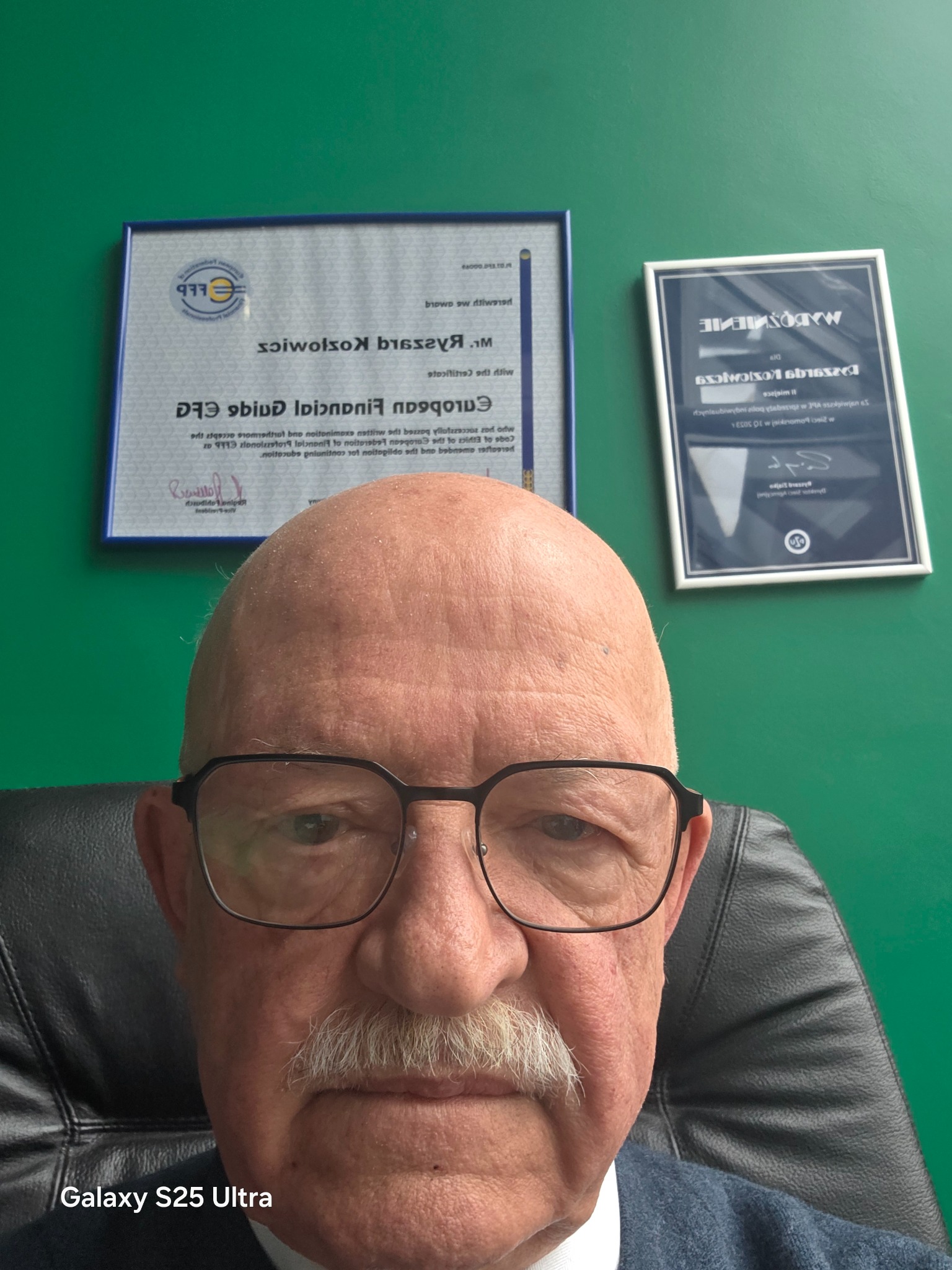 Portret mężczyzny w okularach na tle zielonej ściany z certyfikatami. Widoczne dyplomy EFG i WARDOSNIENIE. Zdjęcie wykonane telefonem Galaxy S25 Ultra.