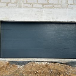 PIENIAK SPÓŁKA Z OGRANICZONĄ ODPOWIEDZIALNOŚCIĄ - Zamknięta, segmentowa brama garażowa w kolorze grafitowym, z poziomymi przetłoczeniami, zamontowana w surowym otworze budynku z bloczków betonowych. Widoczna folia kubełkowa.