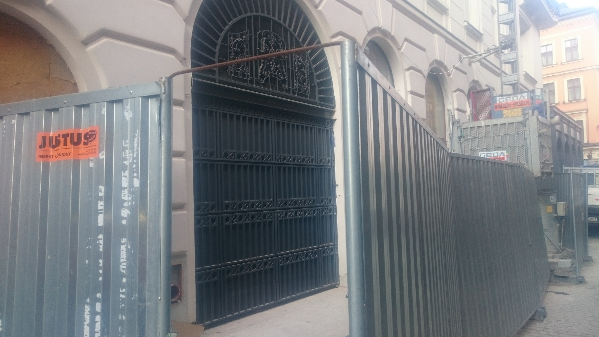 Metalowa brama z ozdobnym łukiem, otoczona tymczasowym ogrodzeniem budowlanym. Widoczny budynek w tle i podnośnik budowlany.