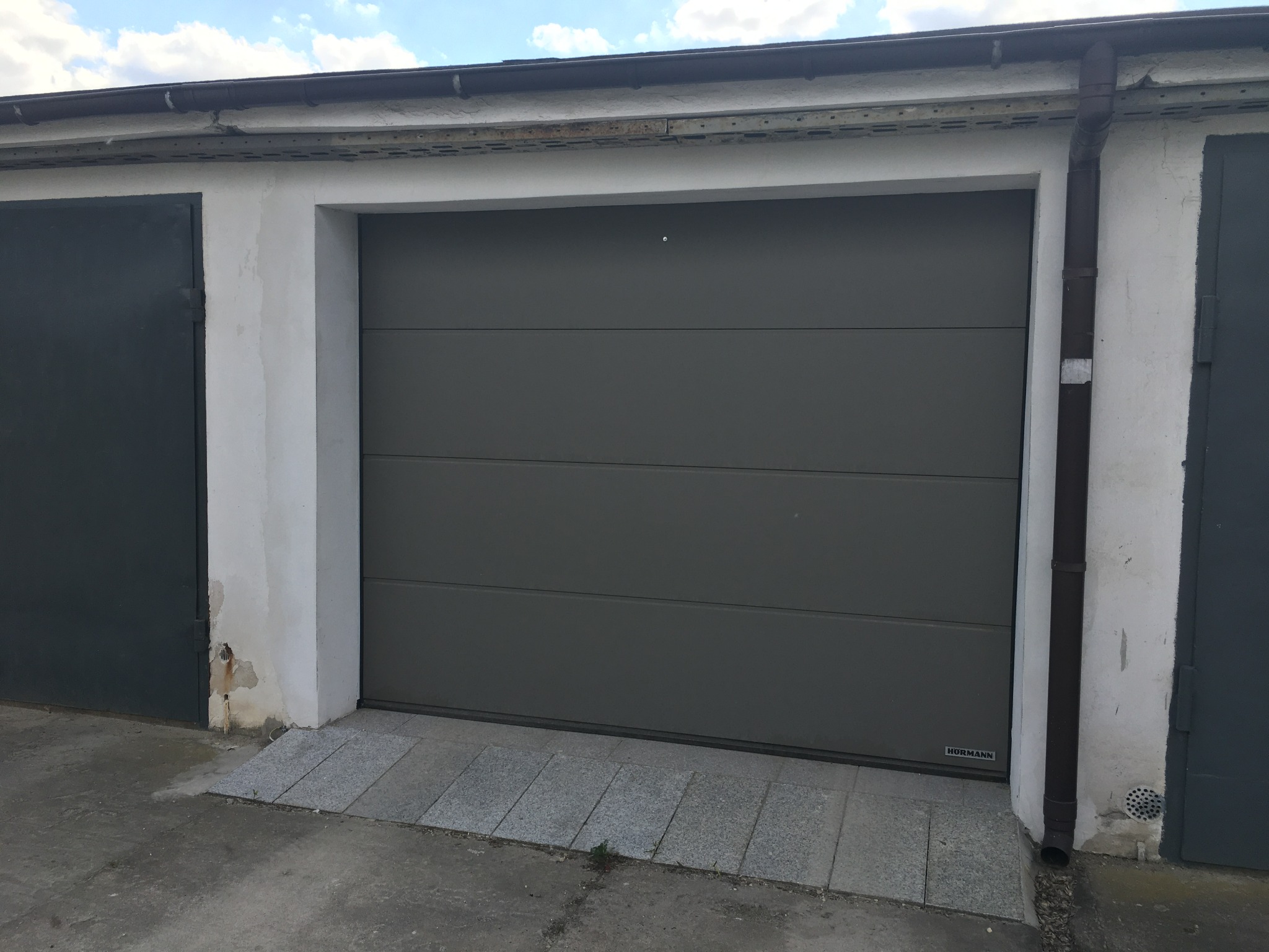 Szara segmentowa brama garażowa marki Hormann, z widocznymi poziomymi panelami, zamontowana w białej wnęce garażu z kostką brukową przed nią. Widoczne rynny i sąsiednie garaże.