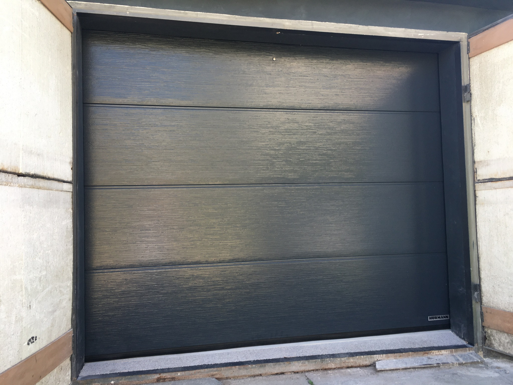 Zamknięta, segmentowa brama garażowa w kolorze antracytowym, z poziomymi przetłoczeniami i widocznym logo producenta w prawym dolnym rogu. Brama osadzona w betonowym otworze.