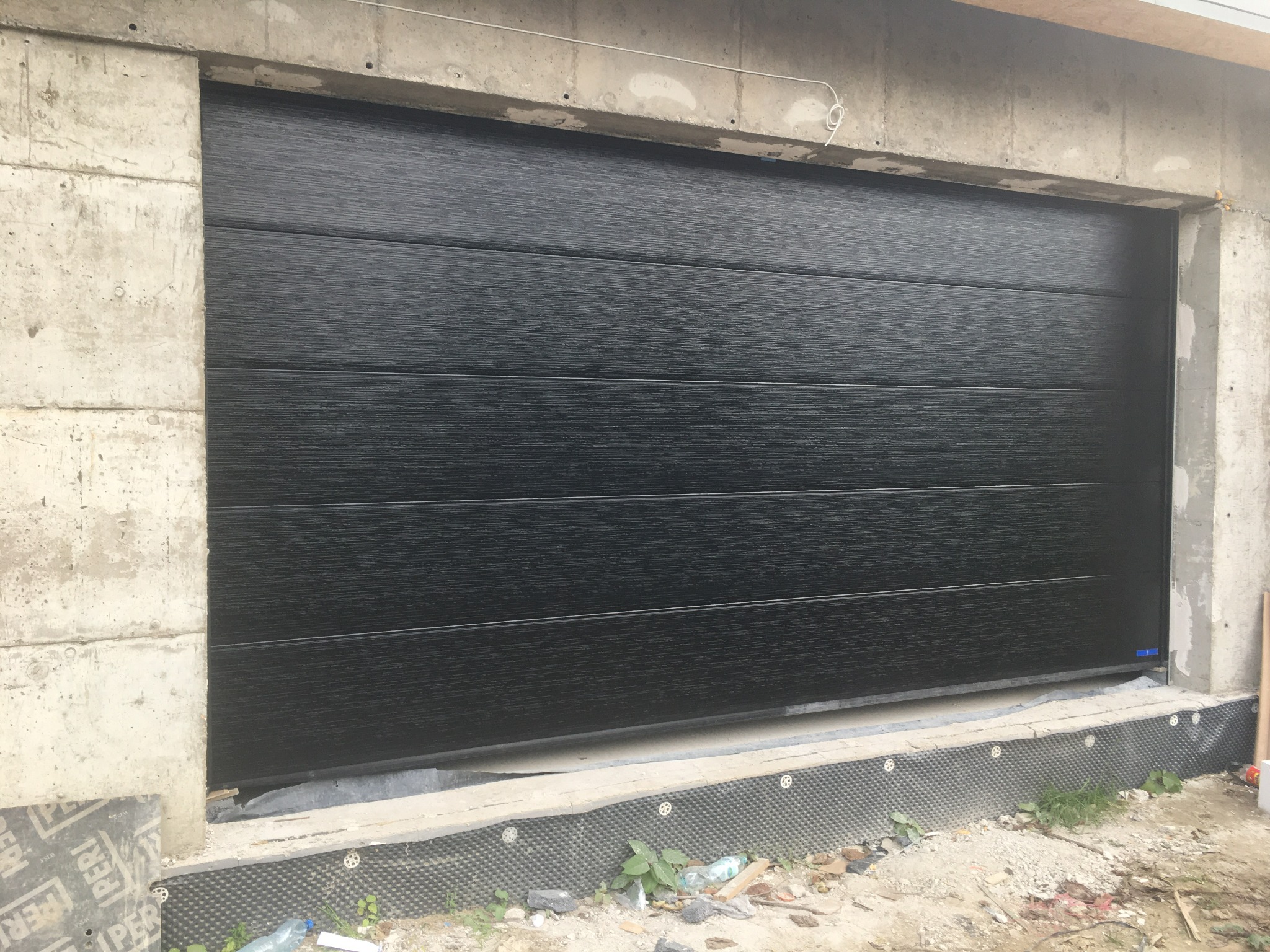 Zamknięta, czarna brama garażowa segmentowa z poziomymi przetłoczeniami, zamontowana w surowym, betonowym otworze budynku w trakcie budowy.