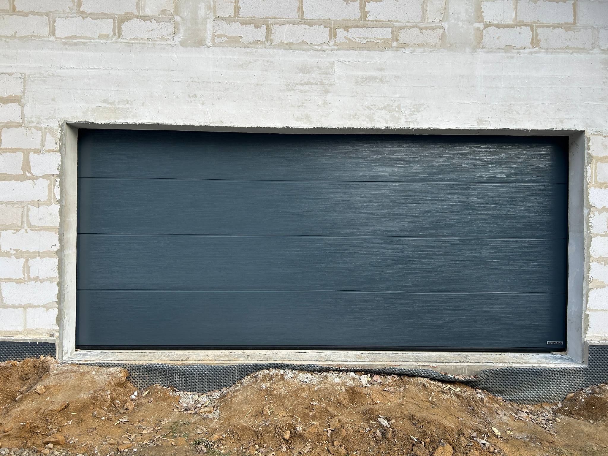 Zamknięta, segmentowa brama garażowa w kolorze grafitowym, z poziomymi przetłoczeniami, zamontowana w surowym otworze budynku z bloczków betonowych. Widoczna folia kubełkowa.