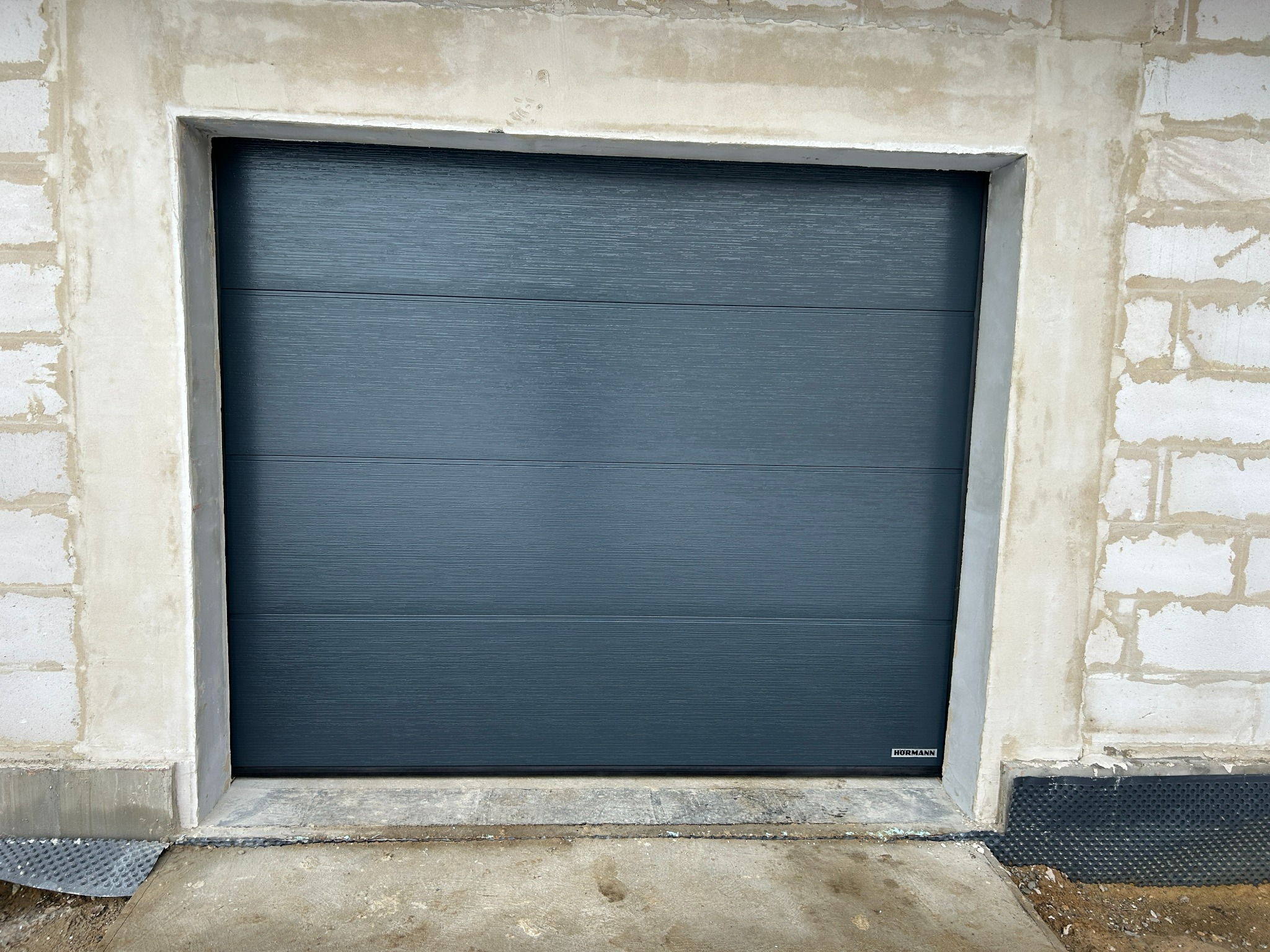 Zamknięta, segmentowa brama garażowa w kolorze grafitowym, z poziomymi przetłoczeniami, zamontowana w surowym otworze budynku z betonu komórkowego.