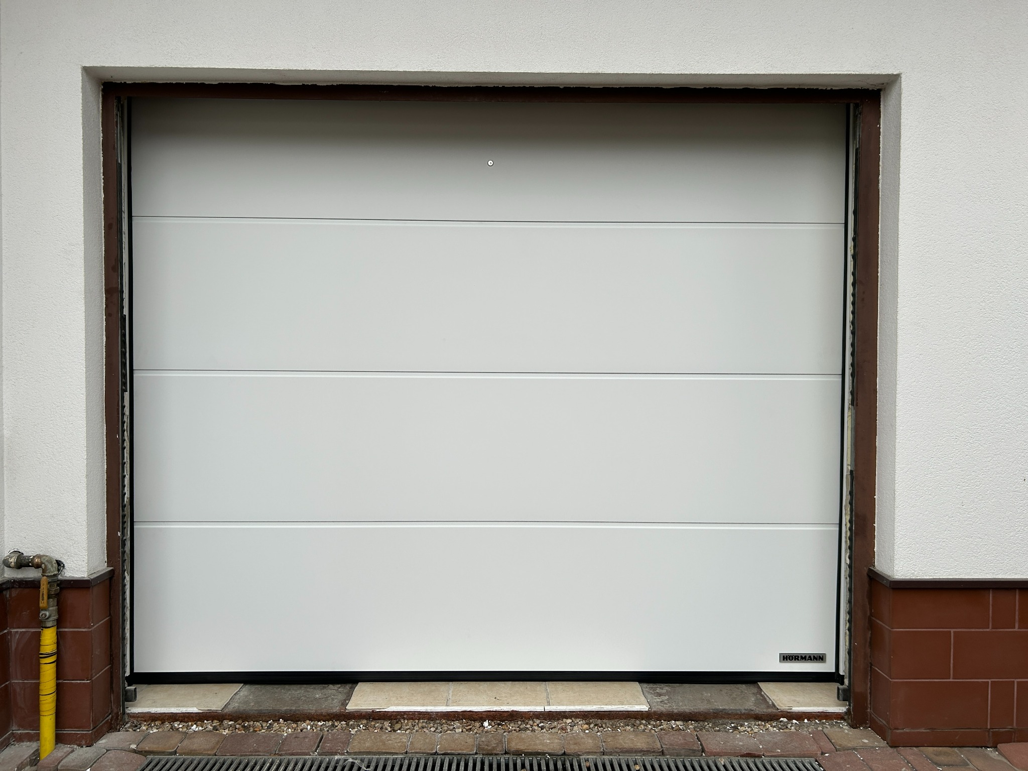 Biała segmentowa brama garażowa marki Hormann, widoczna z zewnątrz, z brązową ramą i elementami wykończeniowymi. Na dole widoczny odpływ liniowy.