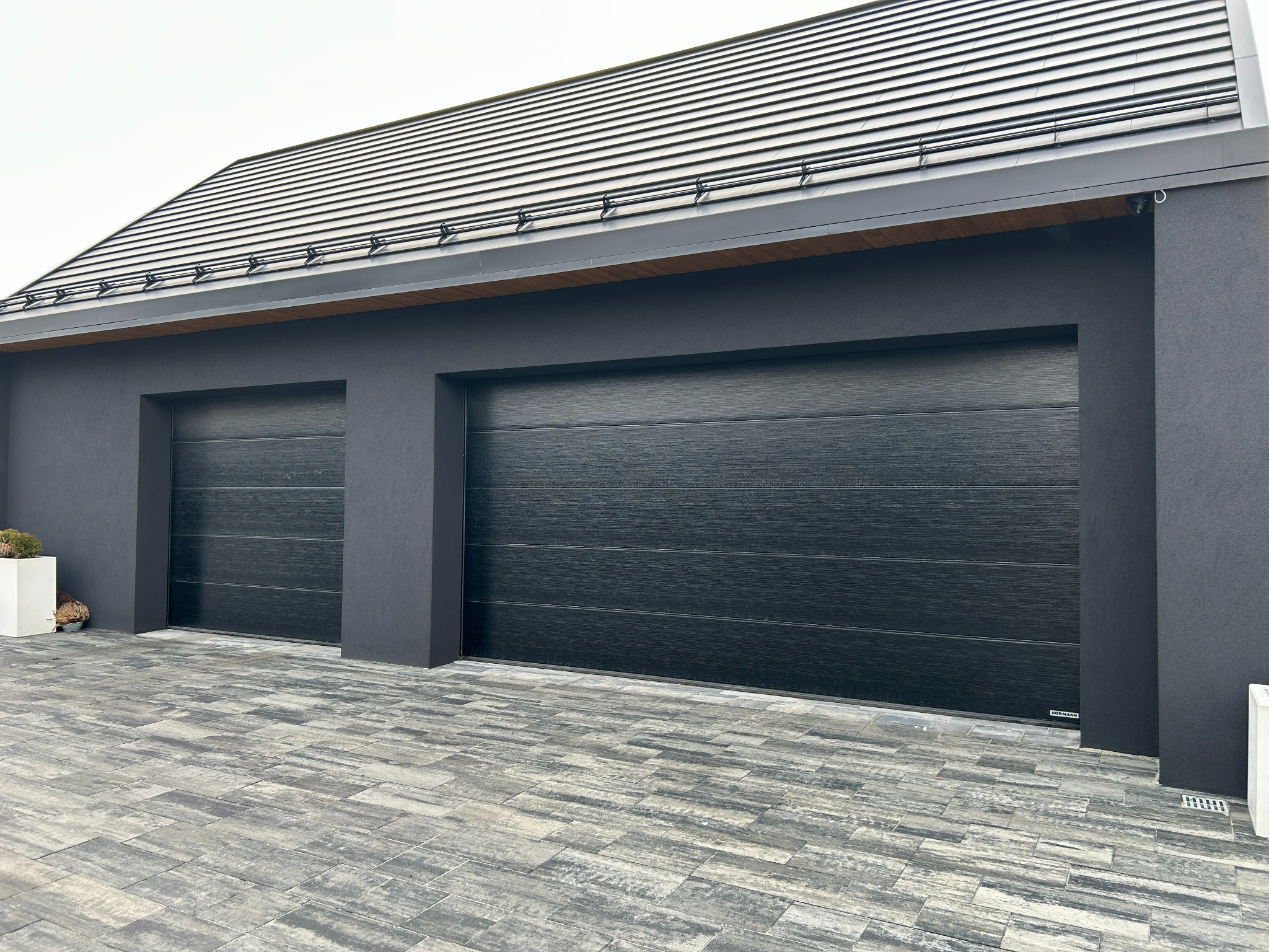 Nowoczesna, czarna brama garażowa segmentowa z poziomymi przetłoczeniami, zamontowana w ciemnoszarym budynku z kostką brukową. Widoczny fragment dachu z szarą dachówką.