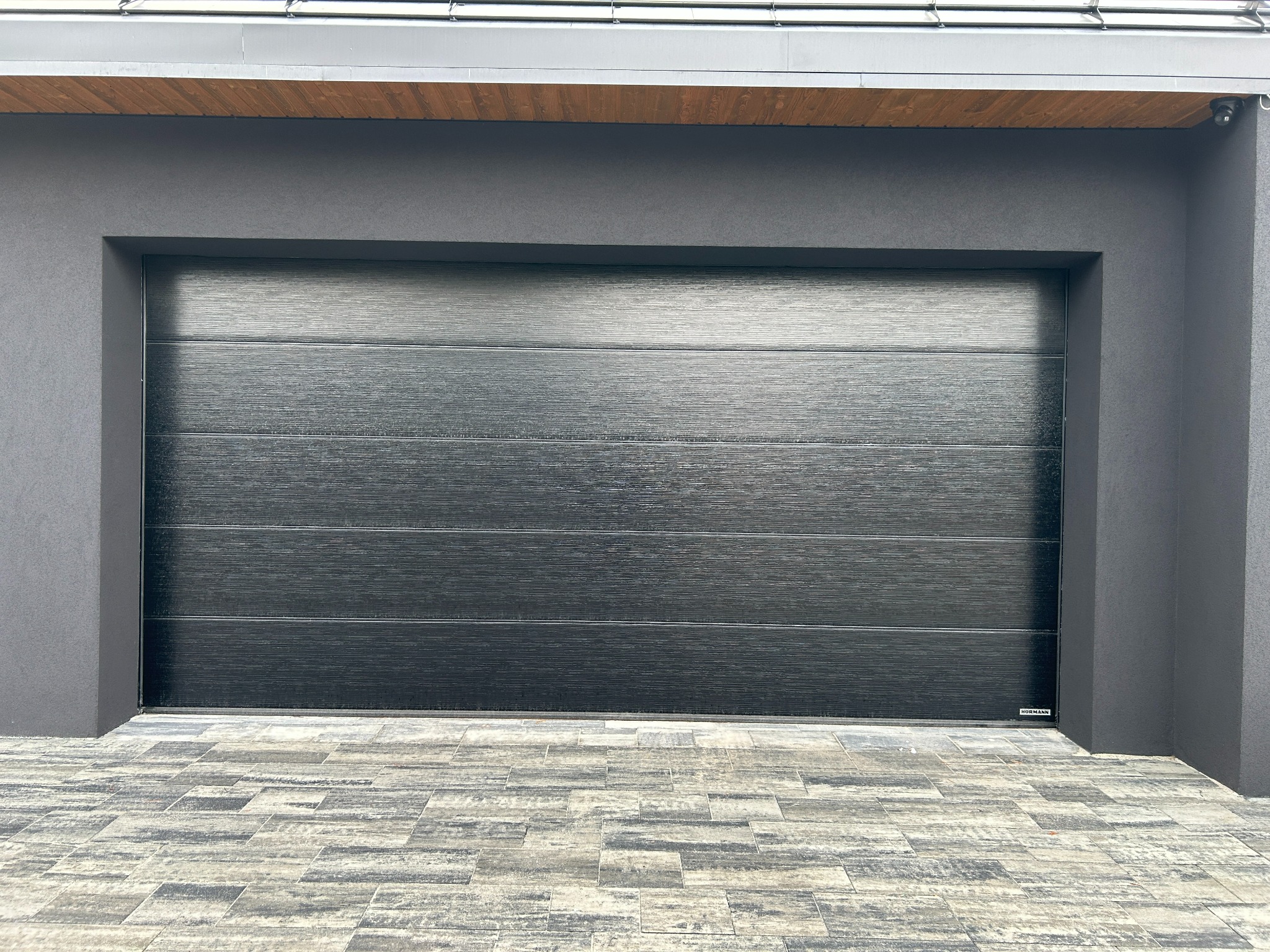 Czarna, segmentowa brama garażowa z poziomymi przetłoczeniami, zamontowana w szarej elewacji budynku. Widoczna kostka brukowa przed bramą i fragment dachu.