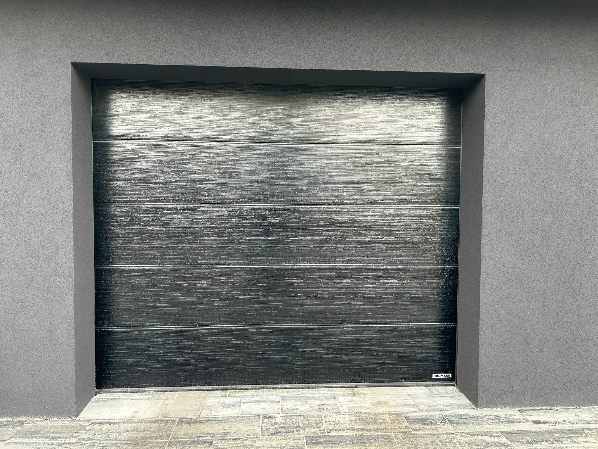 Zamknięta, pozioma, segmentowa brama garażowa w odcieniach szarości, z delikatną fakturą drewna, wkomponowana w elewację budynku. Widoczny fragment chodnika z kostki.