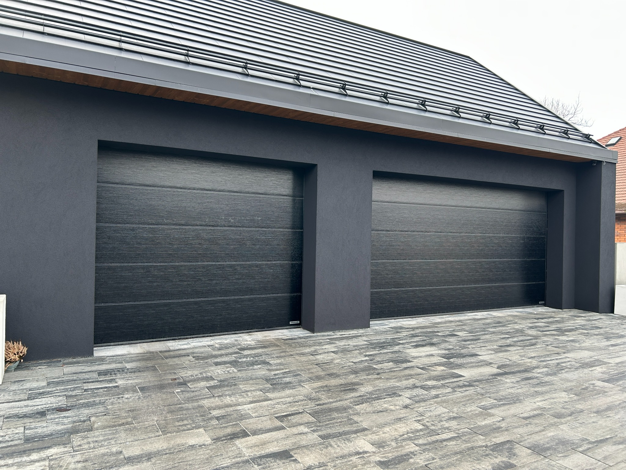 Nowoczesny garaż z dwiema segmentowymi bramami w kolorze antracytu, z wyraźną fakturą drewna. Elewacja budynku w ciemnym odcieniu szarości, kostka brukowa w odcieniach szarości.