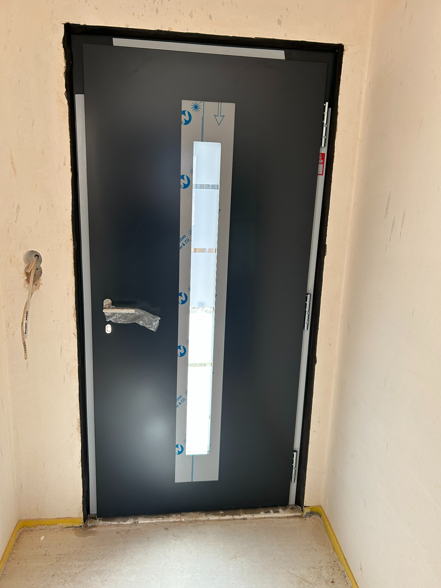 Ciemne drzwi zewnętrzne z długim, wąskim przeszkleniem, zabezpieczone folią ochronną, zamontowane w surowym otworze ściennym. Widoczne kable elektryczne.