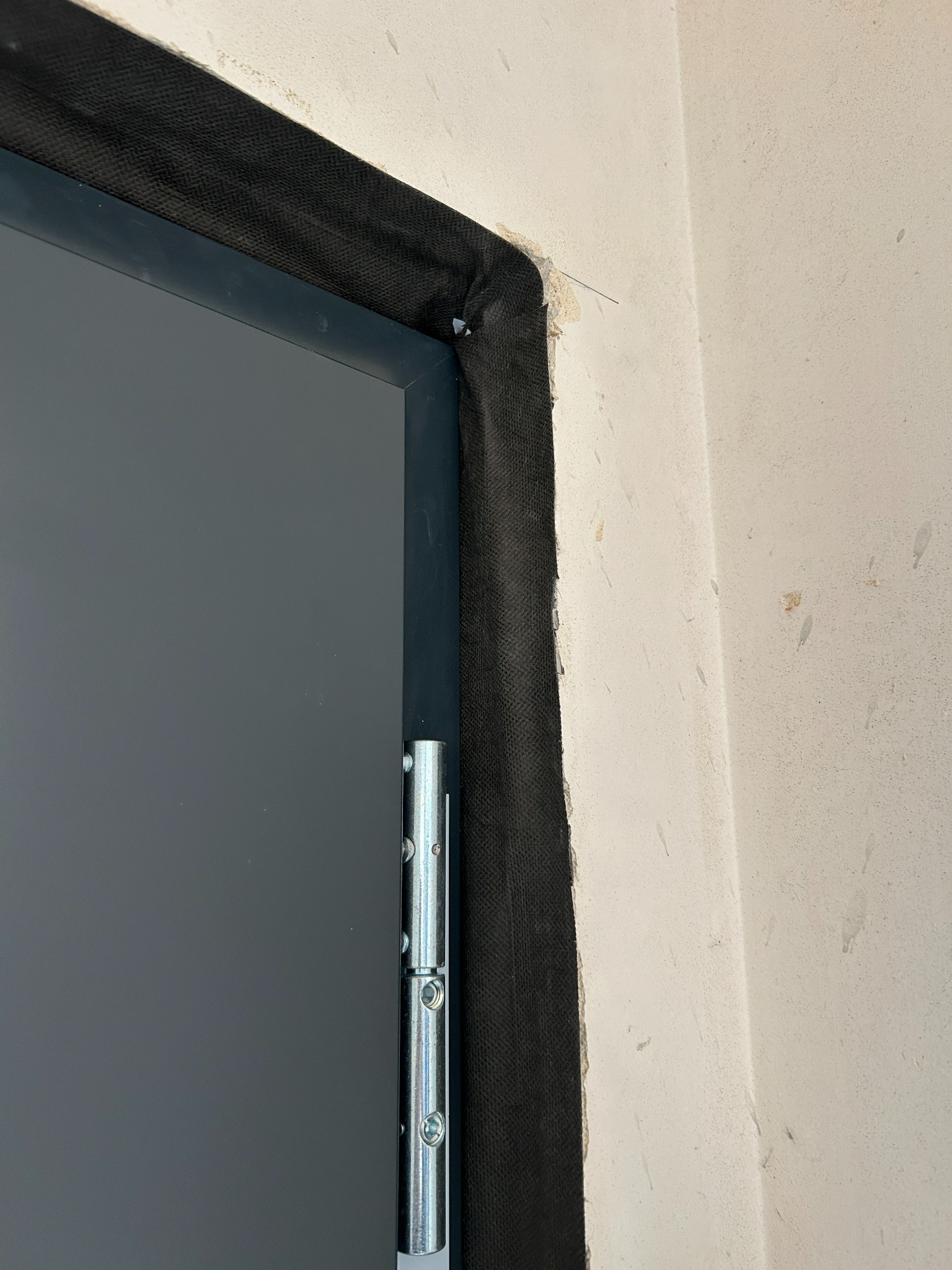 Fragment szarej ościeżnicy z zawiasem, zabezpieczonej czarną taśmą montażową, na tle surowej ściany. Widoczny etap prac wykończeniowych.