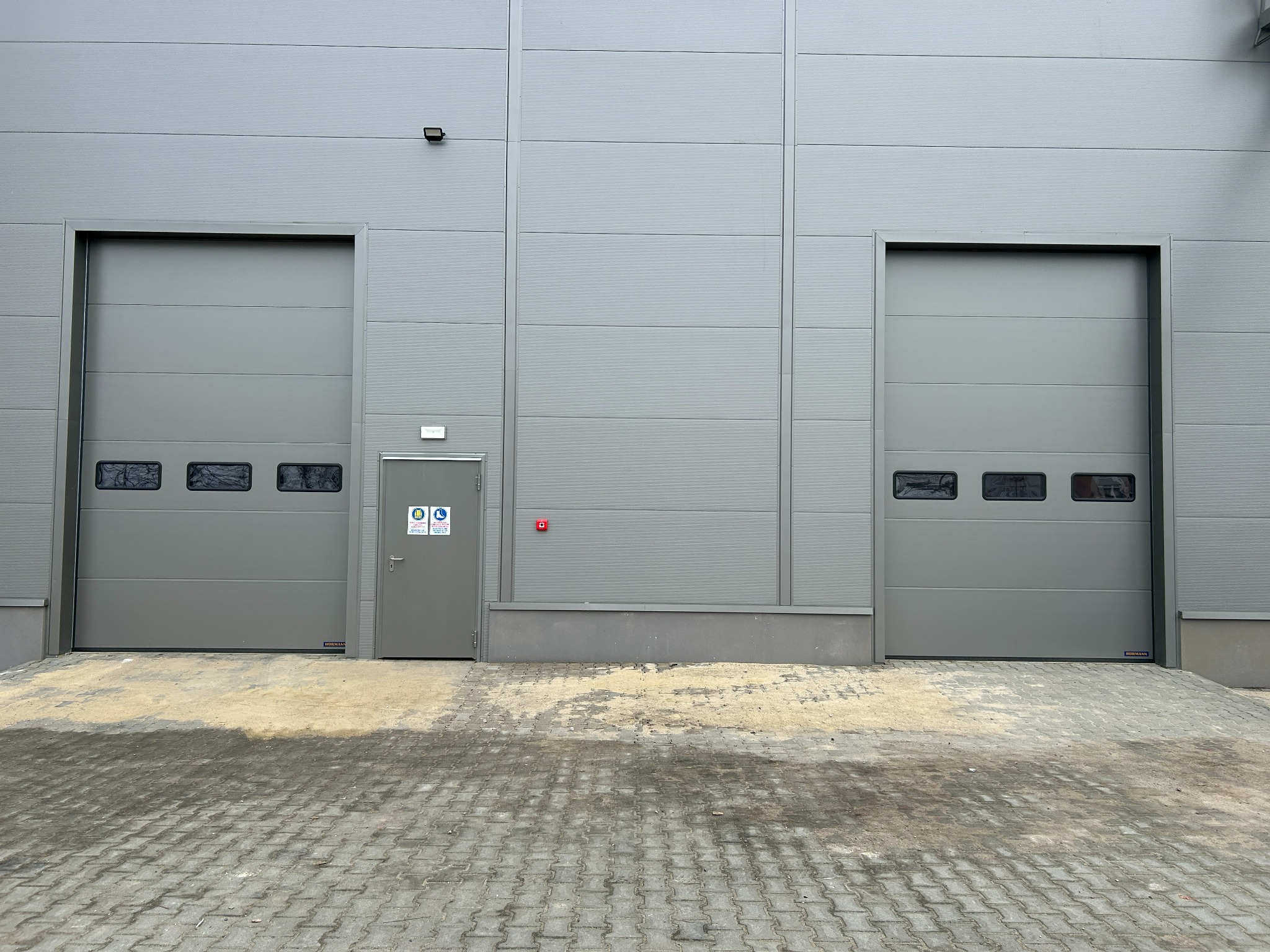 Dwie szare bramy segmentowe z oknami i szare drzwi wejściowe w hali przemysłowej. Widoczne oznaczenia BHP na drzwiach i kostka brukowa przed budynkiem.