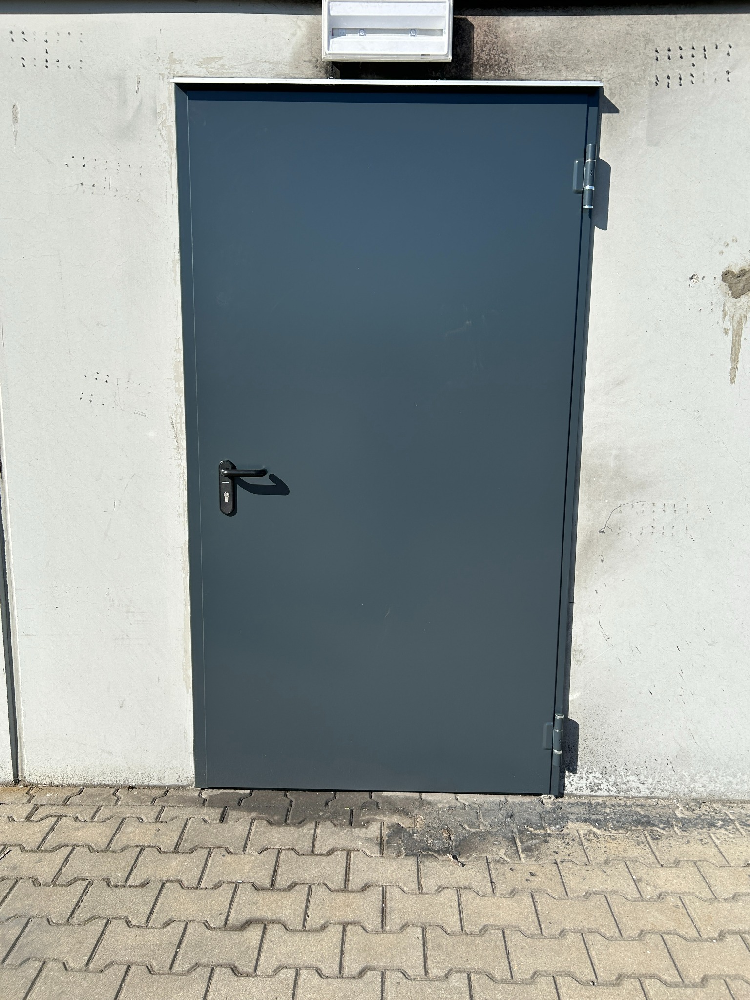 Szara, metalowa brama garażowa z czarną klamką, zamontowana w szarej ścianie z widoczną kostką brukową. Widoczne zawiasy i oświetlenie nad bramą.