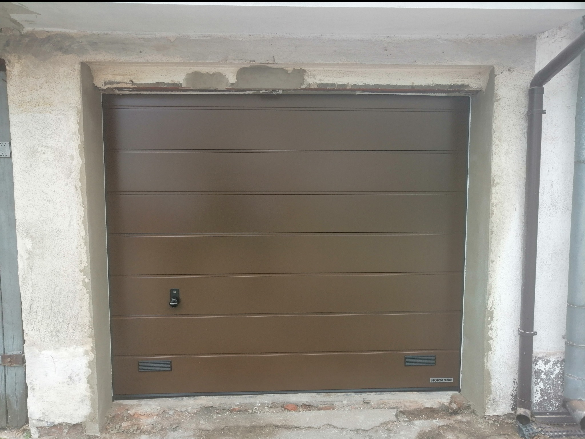 Brązowa brama garażowa segmentowa marki Hormann, zamontowana w surowym otworze garażowym z widocznymi śladami napraw tynkarskich i rynną spustową po prawej stronie.