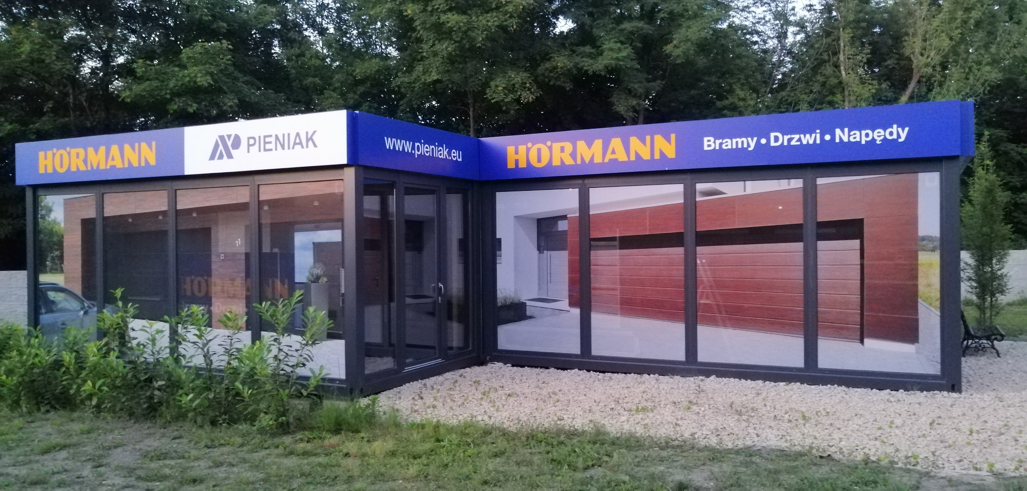 Ekspozycja salonu z bramami garażowymi i drzwiami marki Hormann i Pieniak, otoczona zielenią. Widoczne logo i adres www na górze budynku.