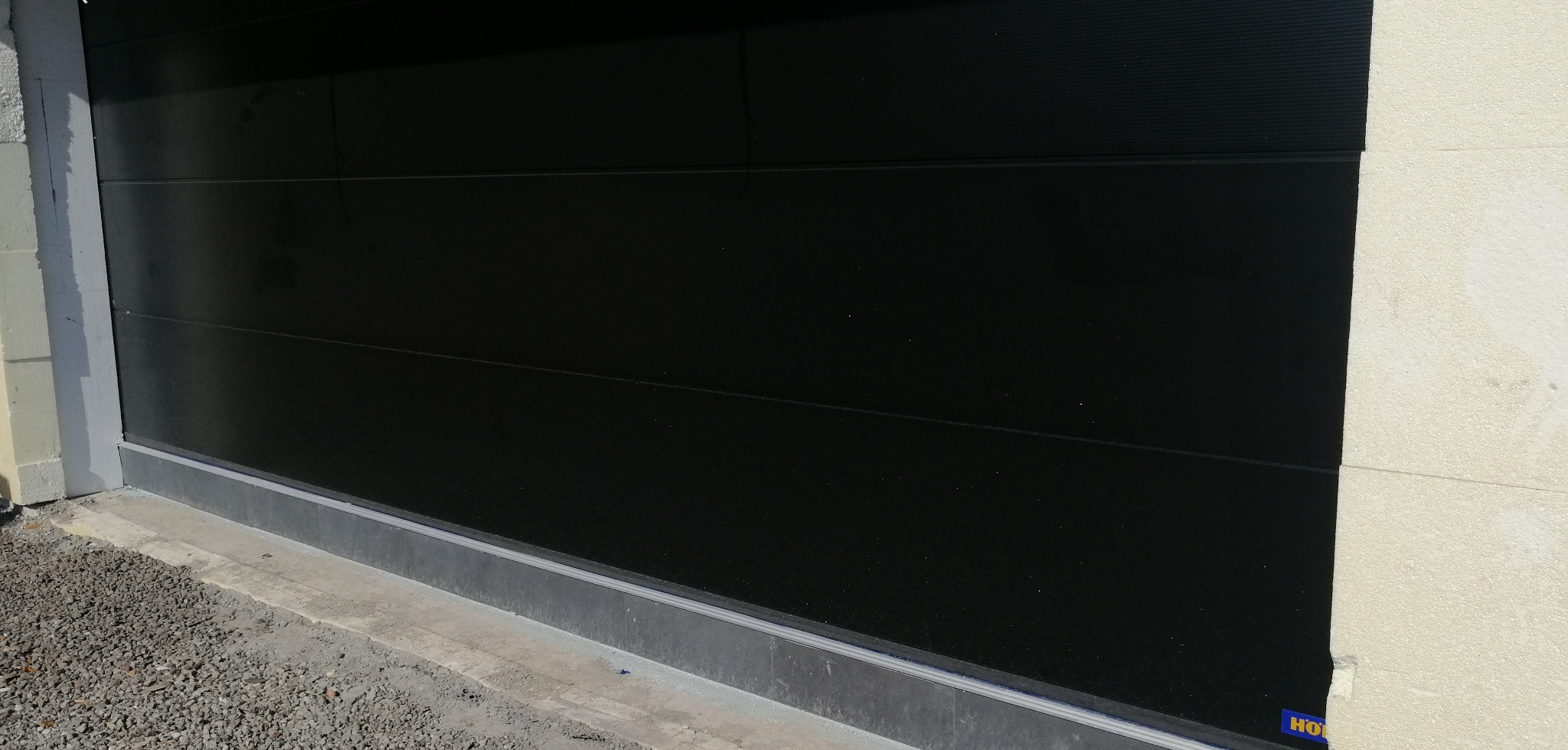 Zamknięta, czarna brama garażowa segmentowa z poziomymi przetłoczeniami, widoczna z bliska, z fragmentem posadzki i ściany. Minimalistyczny design.