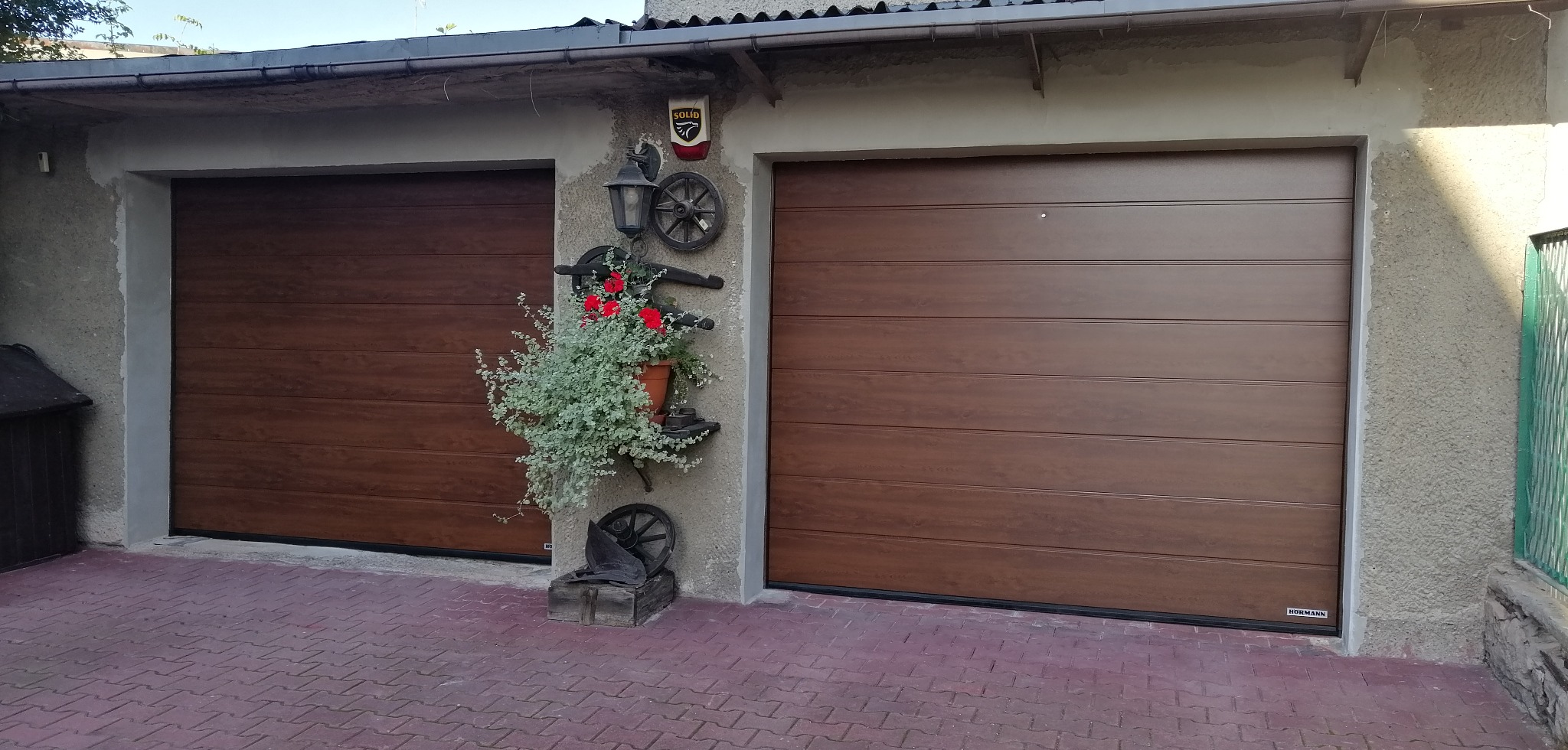 Dwie brązowe, segmentowe bramy garażowe z poziomymi panelami, dekorowane kwiatami i elementami rustykalnymi na ścianie. Widoczny bruk i fragment elewacji.