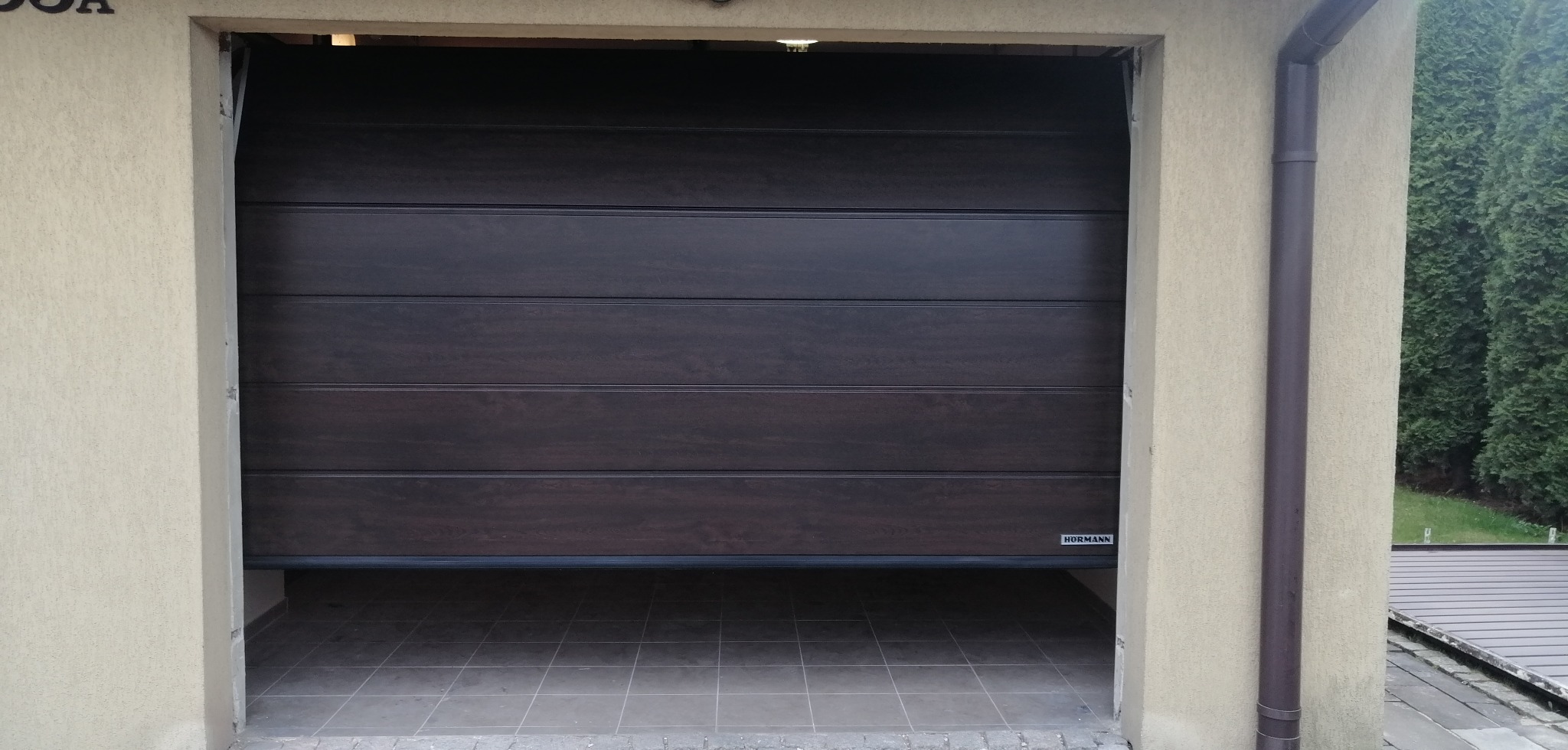 Częściowo otwarta, brązowa brama garażowa segmentowa marki Hormann, widoczna od wewnątrz. Na podłodze szare płytki. Zewnętrzna elewacja budynku w kolorze beżowym.