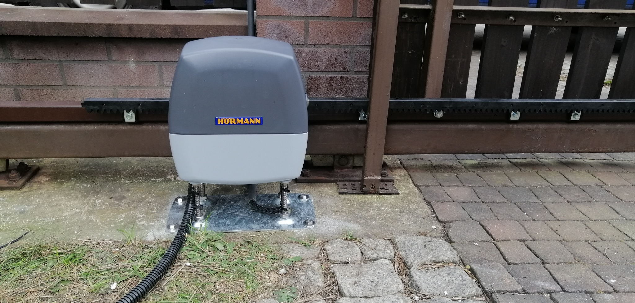 Zamontowany napęd Hormann do bramy przesuwnej, widoczna listwa zębata, podstawa montażowa i przewód zasilający. Tło: ogrodzenie i kostka brukowa.
