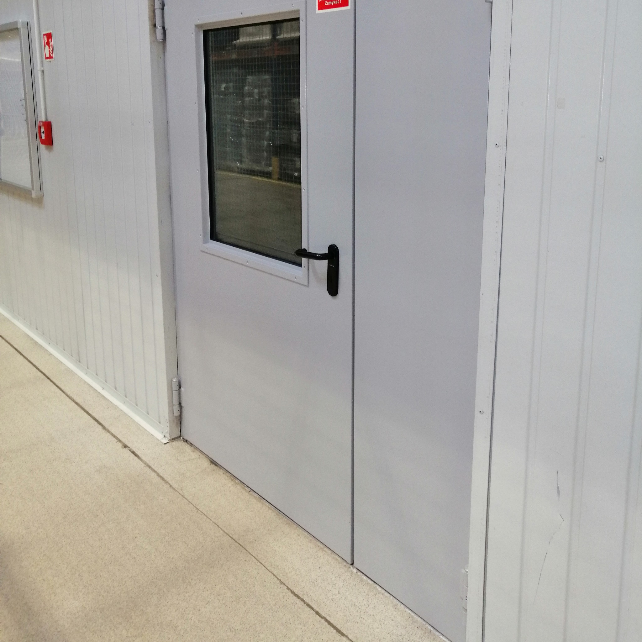 Szare drzwi przemysłowe z małym okienkiem, zamontowane w Będzinie. Widoczna czarna klamka i fragment magazynu przez szybę. Ściany z paneli.