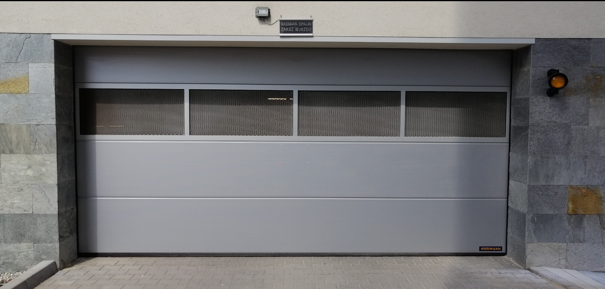 Szara segmentowa brama garażowa z trzema ażurowymi panelami, zamontowana w elewacji wykończonej kamieniem. Widoczny sygnalizator świetlny i tabliczka informacyjna.