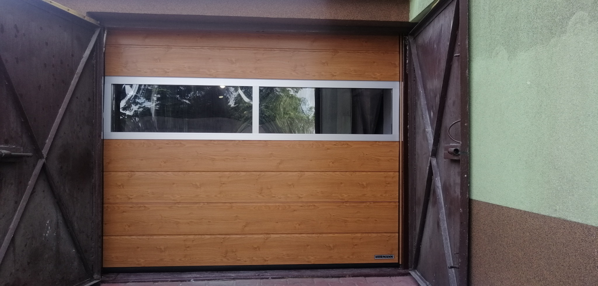 Otwarta brama garażowa segmentowa z poziomymi panelami w odcieniach drewna i oknem z szybą, widok z zewnątrz. Widoczne stare, metalowe drzwi garażu.