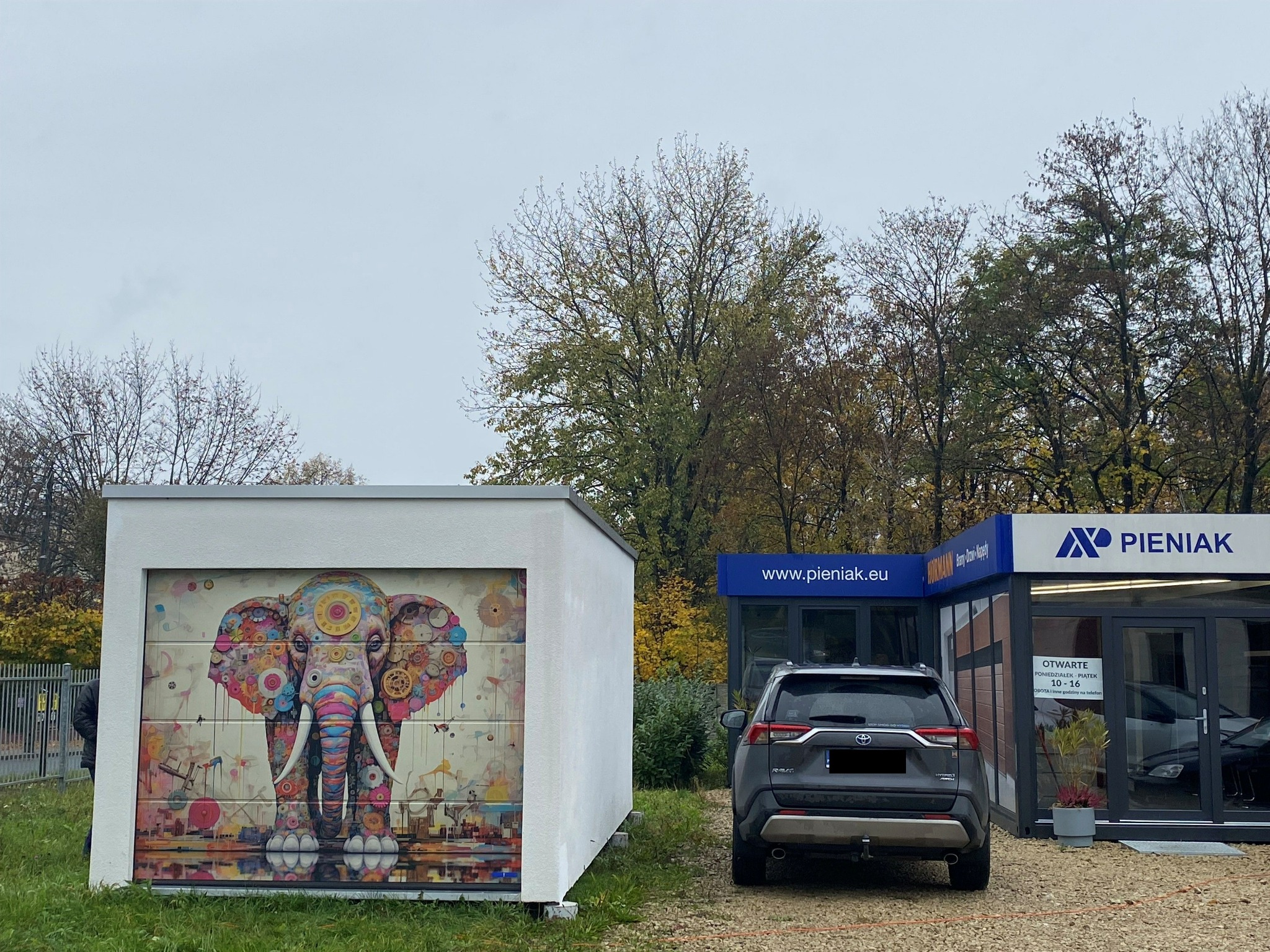Biały garaż z bramą segmentową ozdobioną kolorowym motywem słonia. W tle budynek biurowy i zaparkowany szary SUV. Pochmurny dzień.