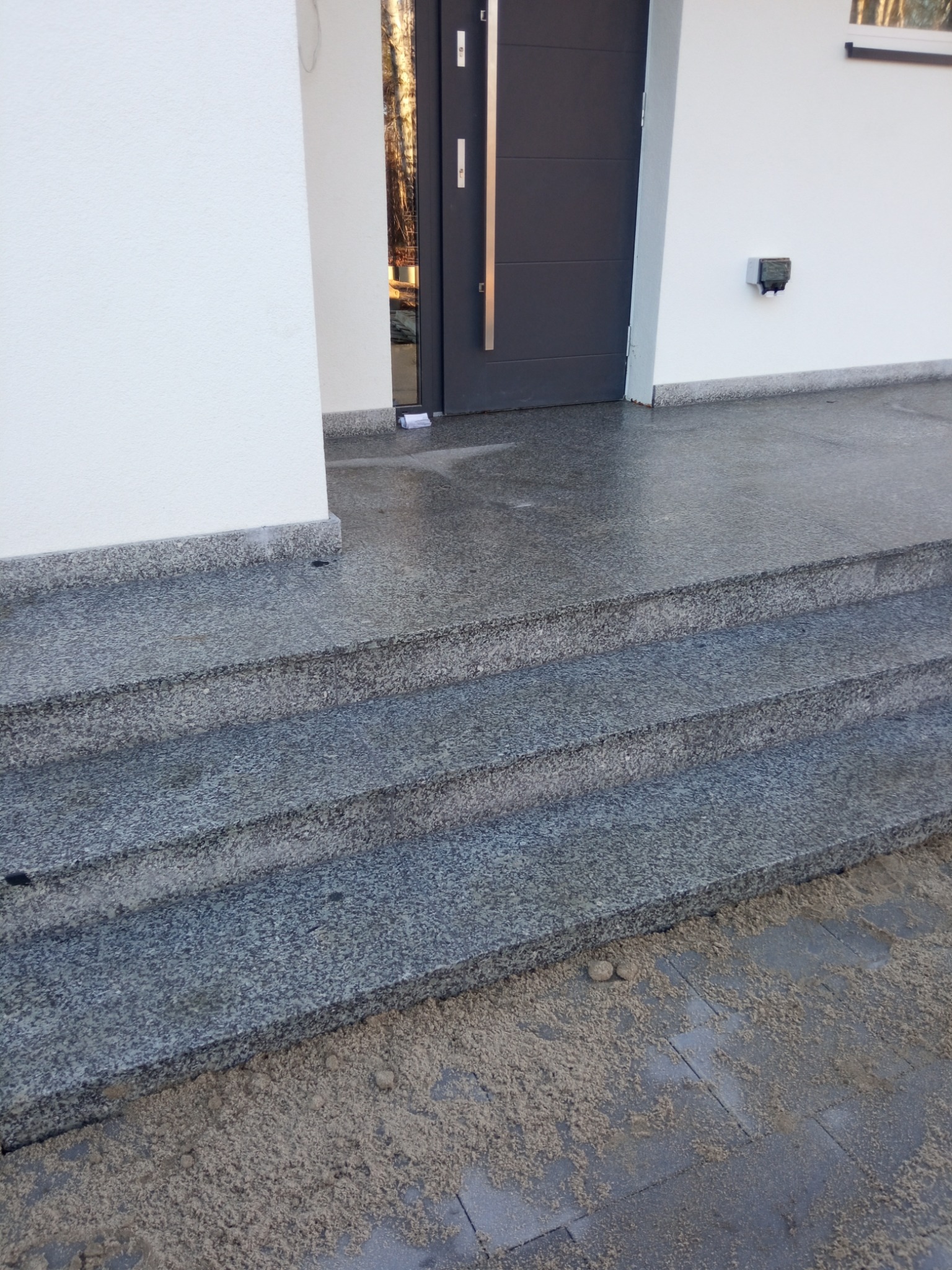 Granitowe schody wejściowe z trzema stopniami prowadzące do nowoczesnego domu z szarymi drzwiami. Widoczne ślady piasku na kostce brukowej.