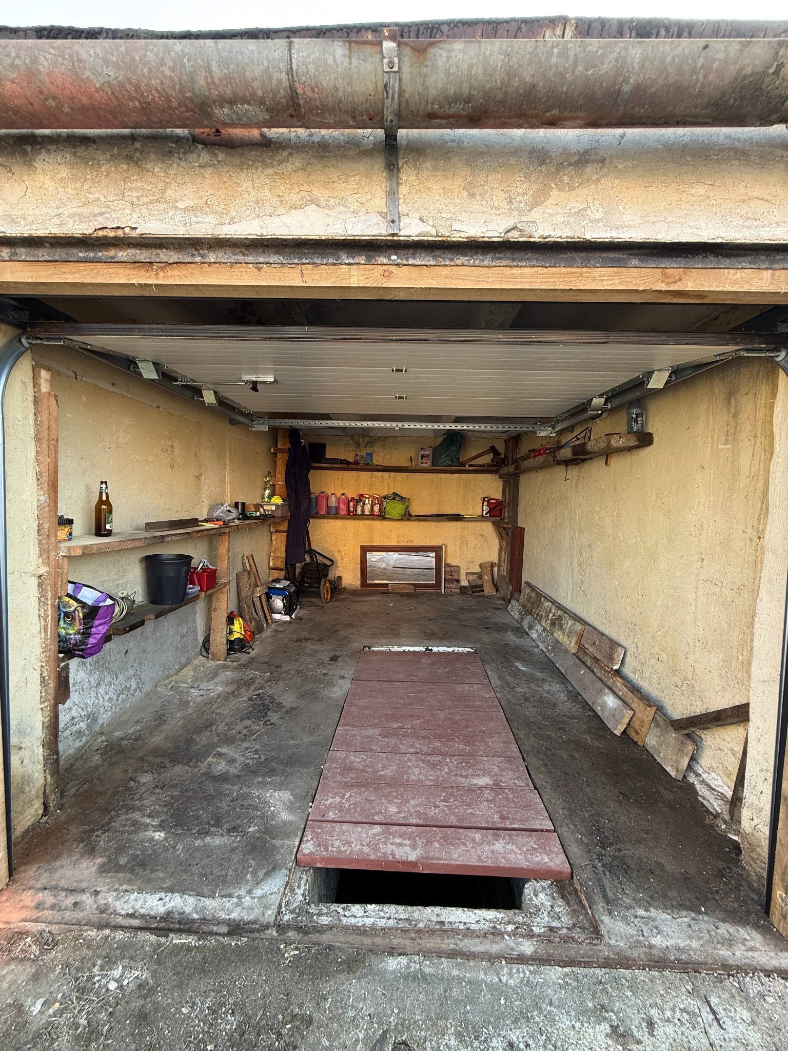 Otwarty garaż z betonową podłogą i drewnianymi półkami, na których stoją różne przedmioty. W tle widoczna osoba w kombinezonie. W podłodze znajduje się drewniana klapa.