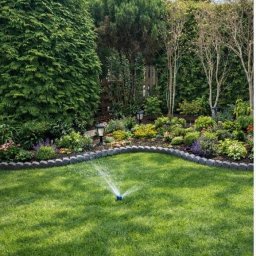 Karol Michalski - Zraszacz rotacyjny nawadnia soczyście zielony trawnik w ogrodzie z ozdobnymi rabatami i tujami. W tle widoczne drzewa i błękitne niebo.