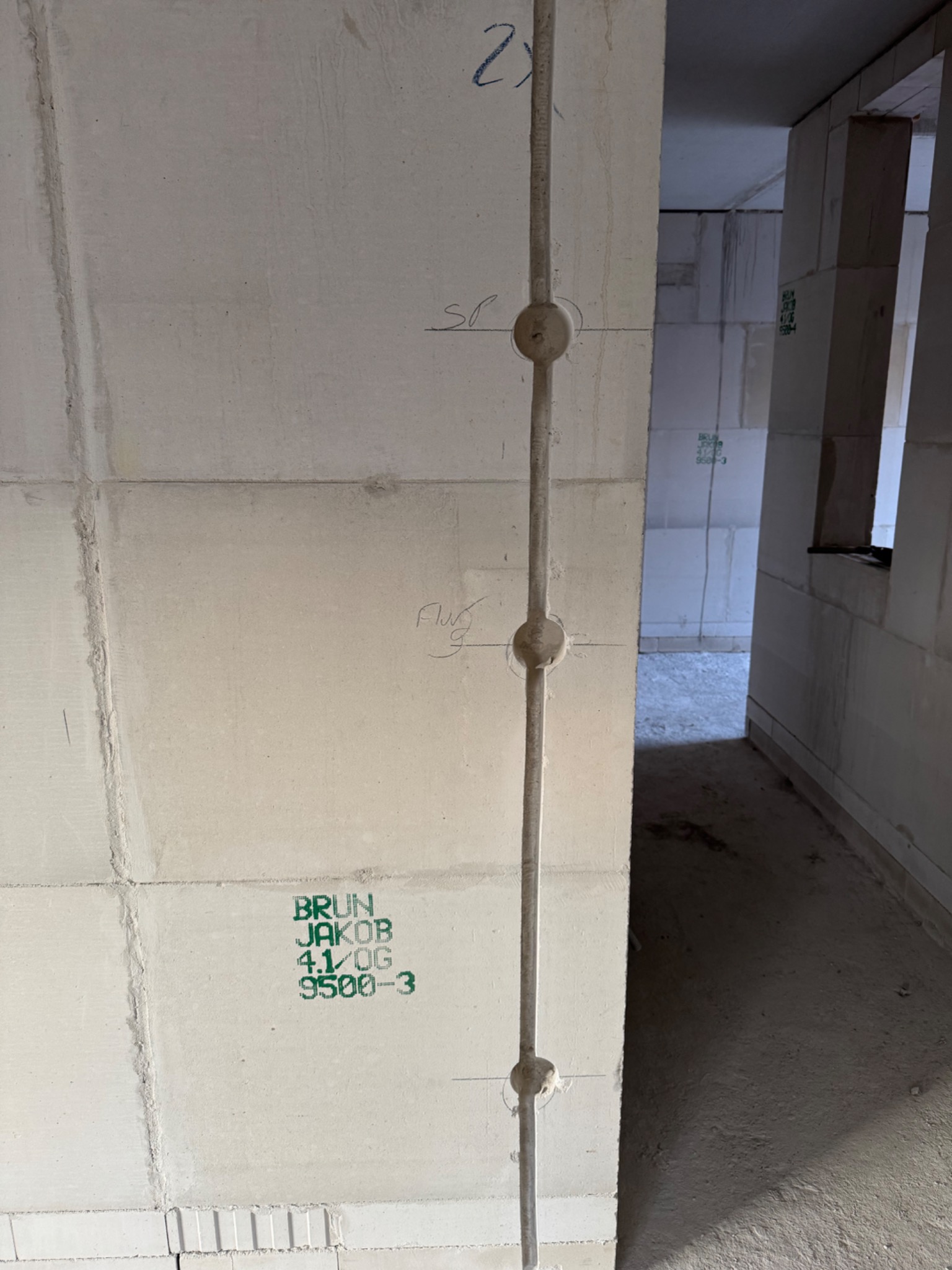 Pionowy przewód elektryczny z puszkami w ścianie z betonu komórkowego, widok na surowy stan budynku w głębi. Instalacja elektryczna w trakcie budowy.