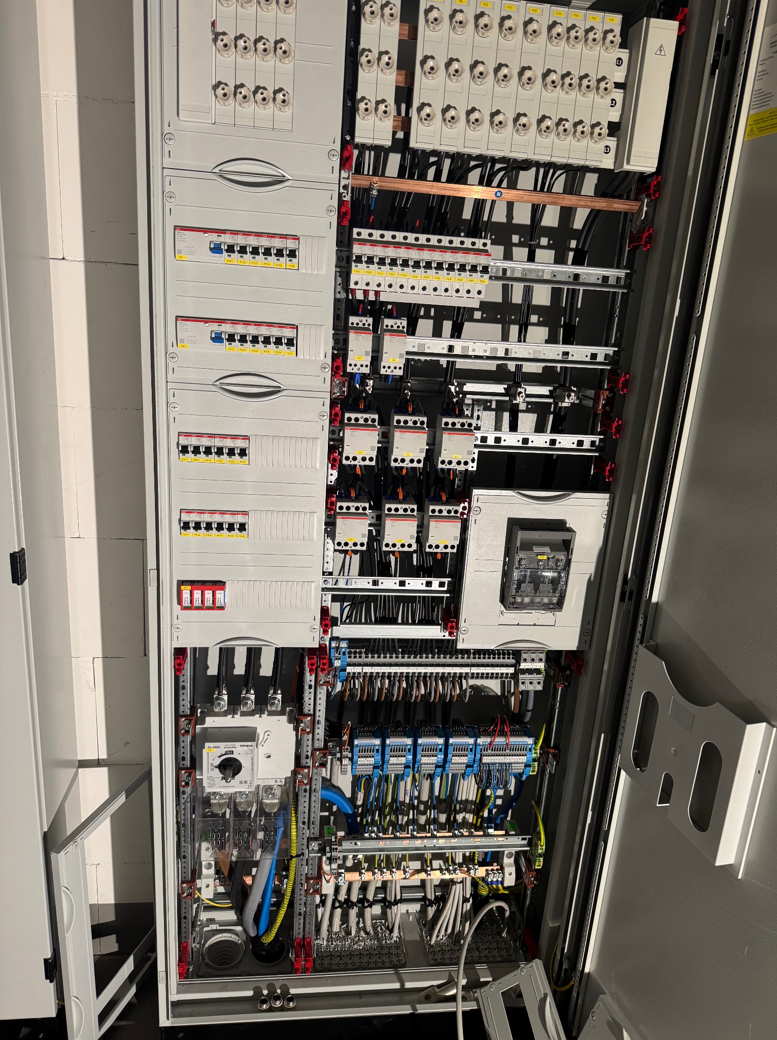 Otwarta szafa rozdzielcza z bezpiecznikami, przewodami i elementami sterującymi. Widoczne liczne kable, listwy zaciskowe i zabezpieczenia. Nowoczesna instalacja elektryczna.