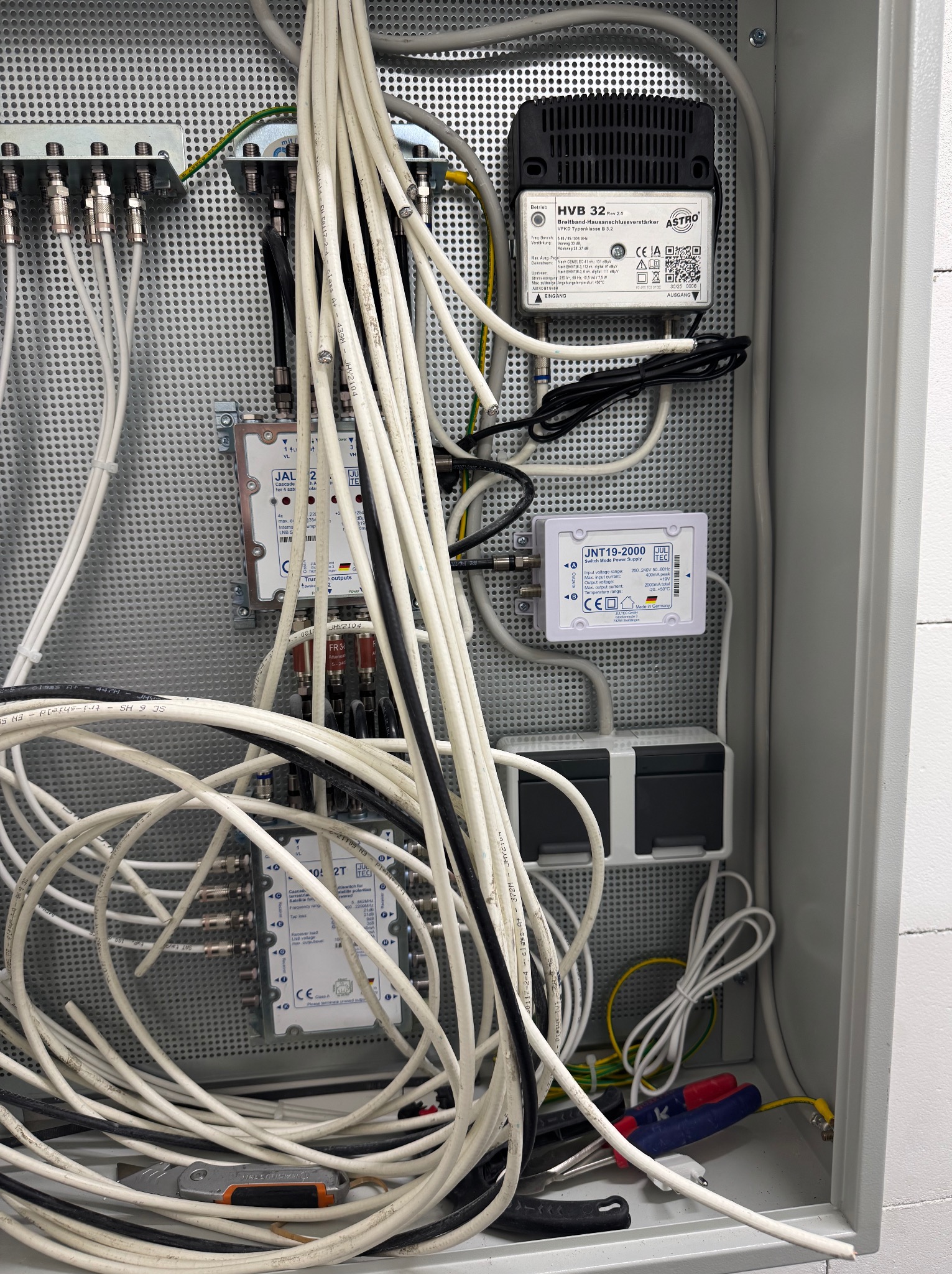 Rozdzielnica z kablami antenowymi i urządzeniami elektronicznymi, widoczne narzędzia. Instalacja antenowa w szarej obudowie z perforowanymi ściankami.
